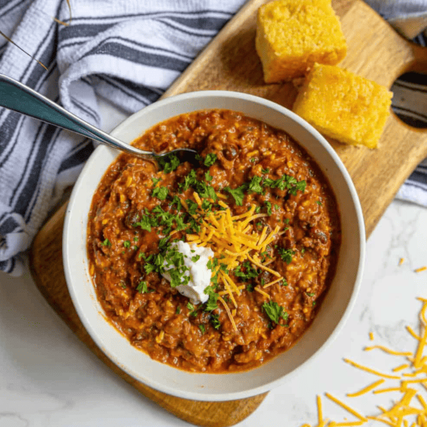 A beefy bean chili with sour cream and cornbread on a cutting board.