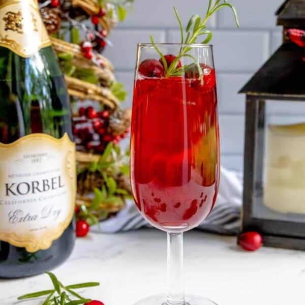 A glass of cranberry champagne cocktail with cranberries and sprigs of rosemary.