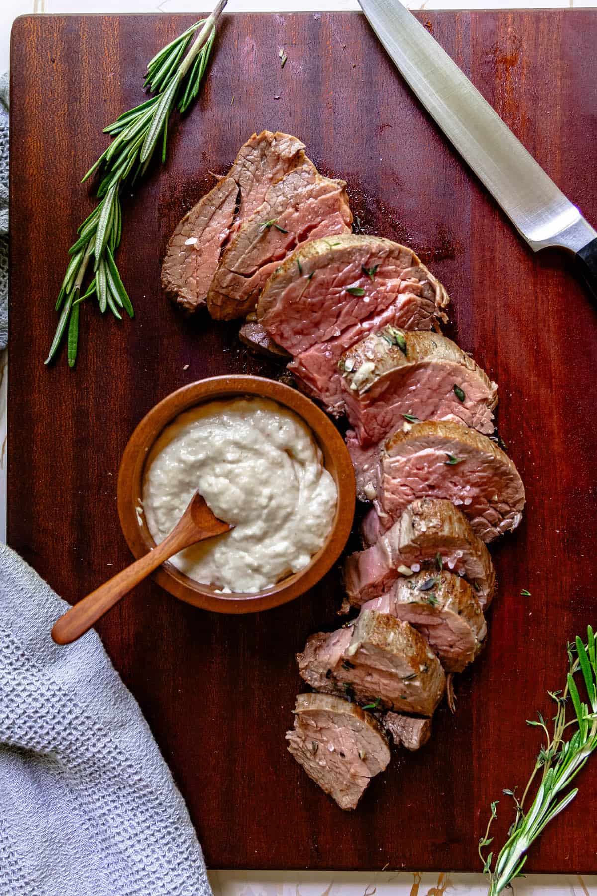 Garlic herb beef tenderloin with sour cream on a cutting board with sprigs of rosemary.