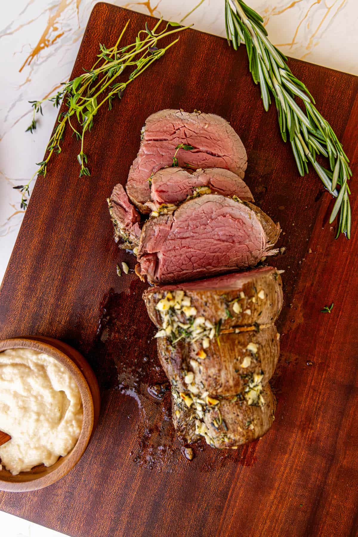 A garlic herb beef tenderloin on a cutting board with a dipping sauce.