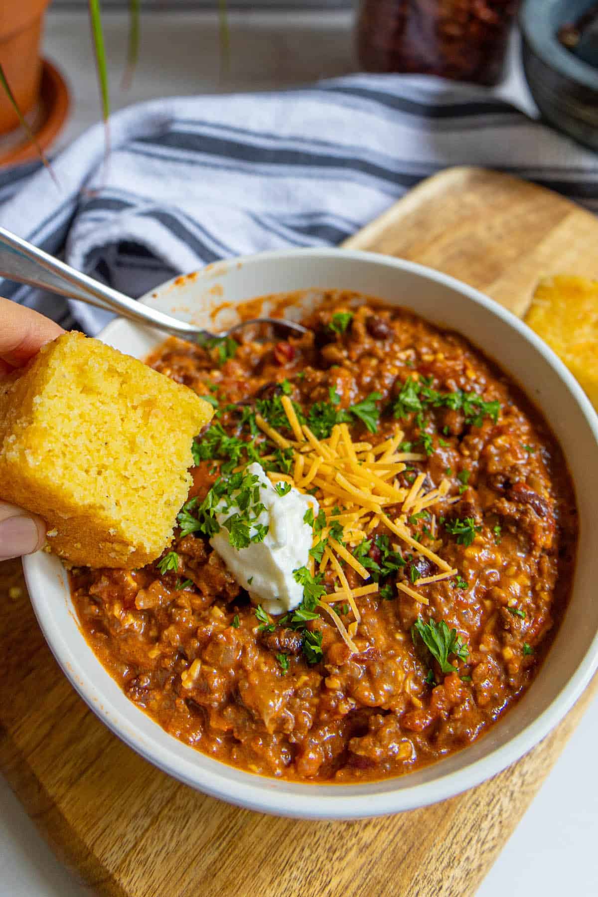 A bowl of chili with cornbread on a cutting board.