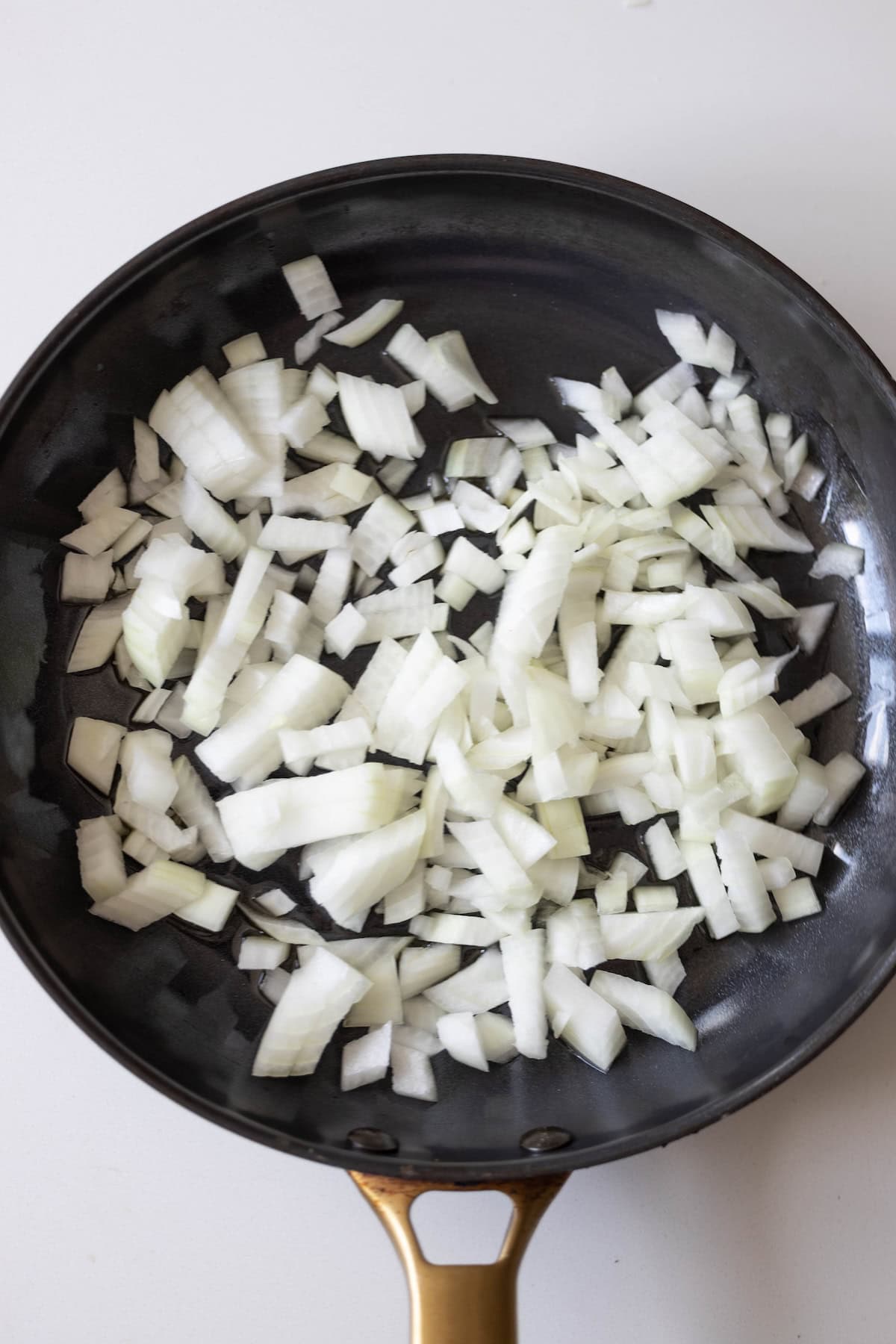 A black frying pan containing chopped raw onions sits on a white surface, ready to start a creamy pepper chicken dinner.