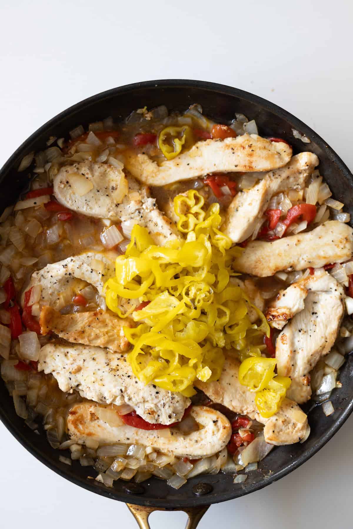 A skillet containing cooked chicken strips, diced onions, chopped tomatoes, and sliced yellow peppers makes the perfect base for a creamy pepper chicken dinner on a white surface.