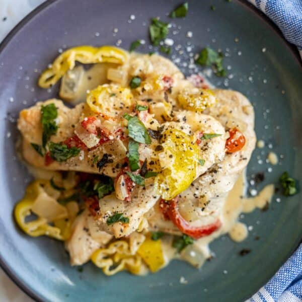 A plate of creamy pepper chicken dinner features cooked chicken breasts topped with luscious sauce, sliced peppers, diced onions, chopped herbs, and cracked black pepper.