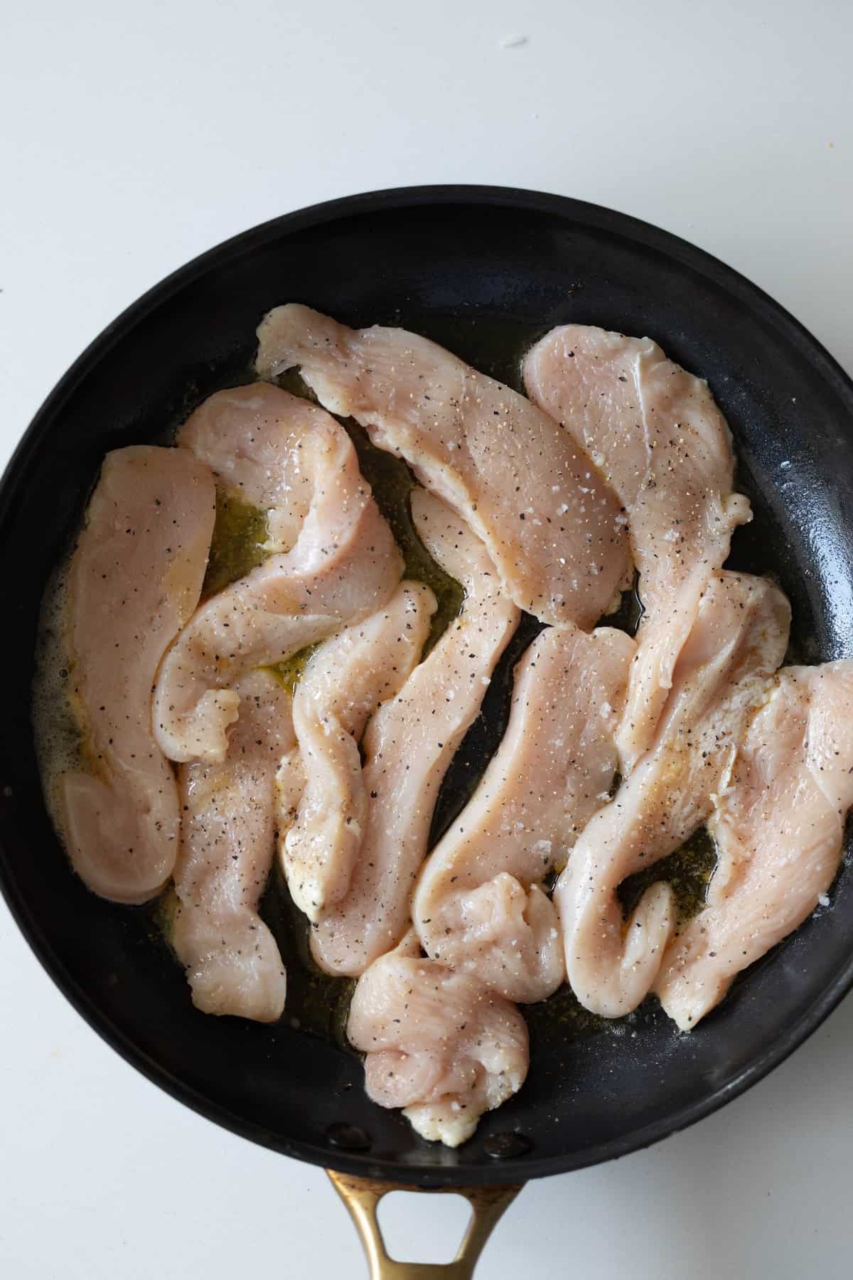 Raw chicken breast strips seasoned with black pepper sizzle in a black skillet with oil on a white surface, perfect for a creamy pepper chicken dinner.