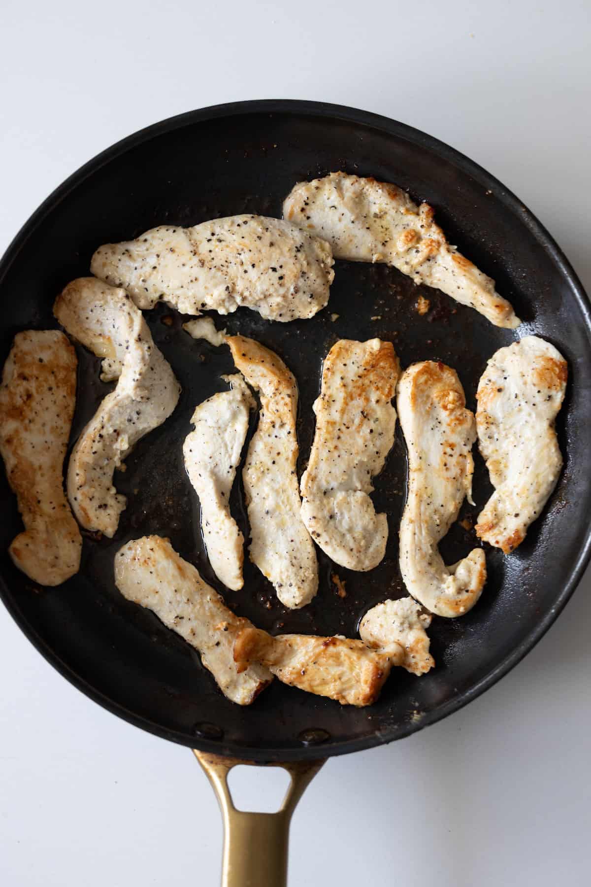 Cooked chicken breast strips seasoned with pepper and salt in a black frying pan on a white surface—perfect for a Creamy Pepperoncini Chicken Skillet dinner.