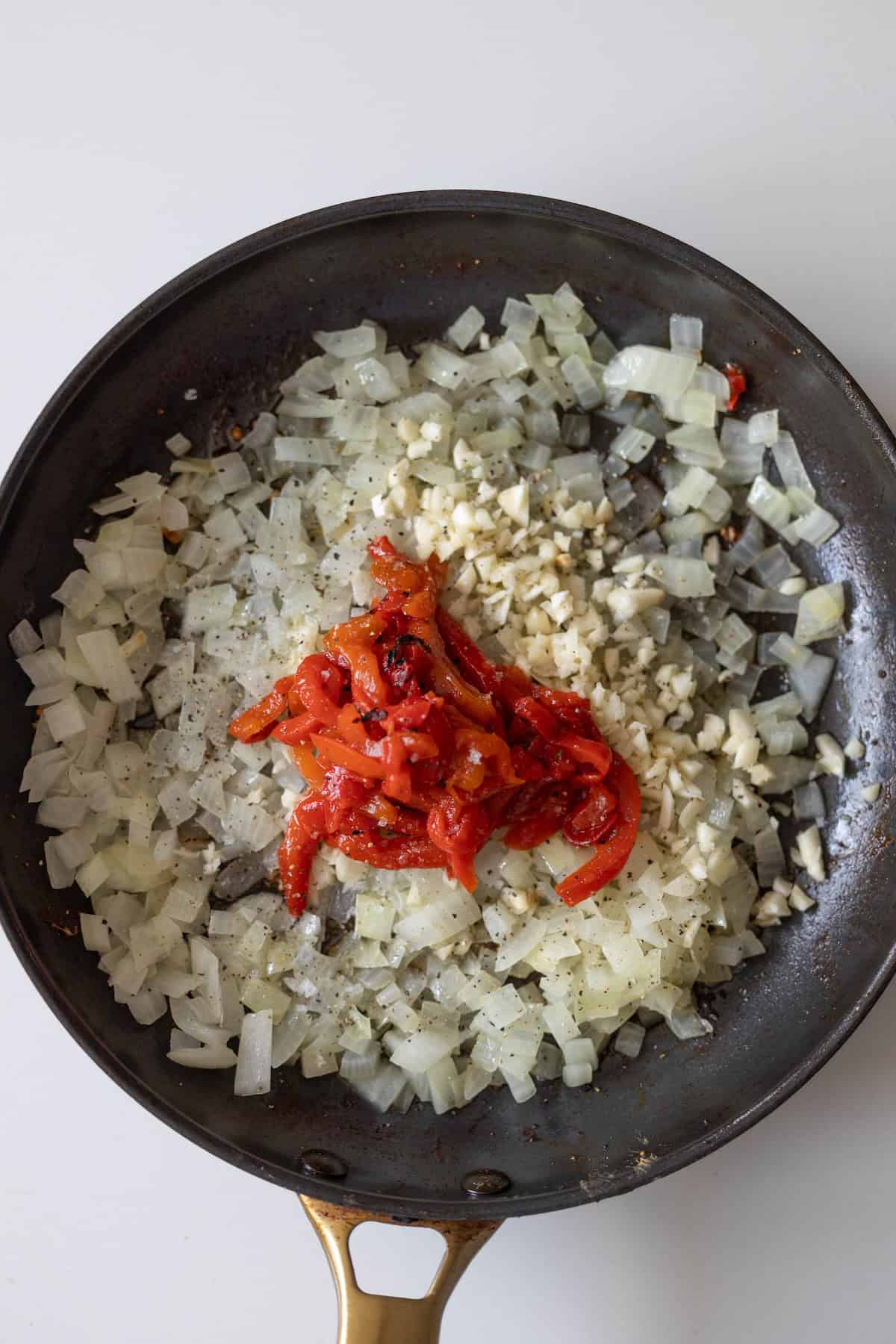 Chopped onions, garlic, and sliced roasted red peppers sizzle in a black skillet with a brass handle, the perfect start to a creamy pepper chicken dinner.