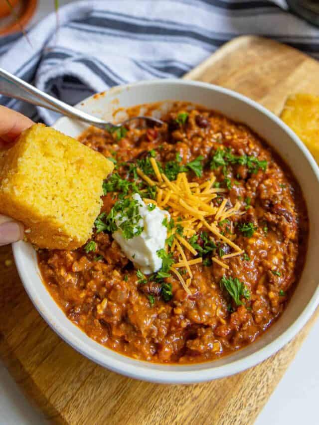 A bowl of chili with cornbread on a cutting board.