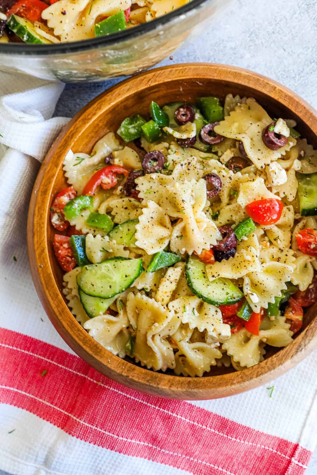 A wooden bowl filled with pasta and vegetables, resembling a Greek salad.