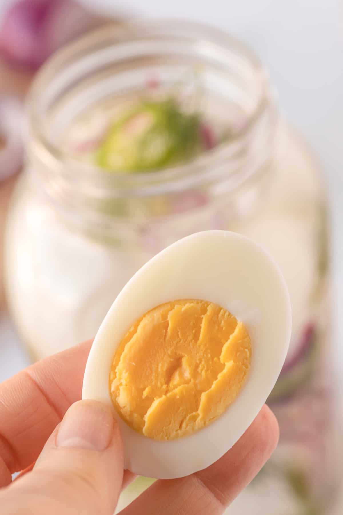 A person holding a hard boiled egg in front of a jar of pickled eggs.