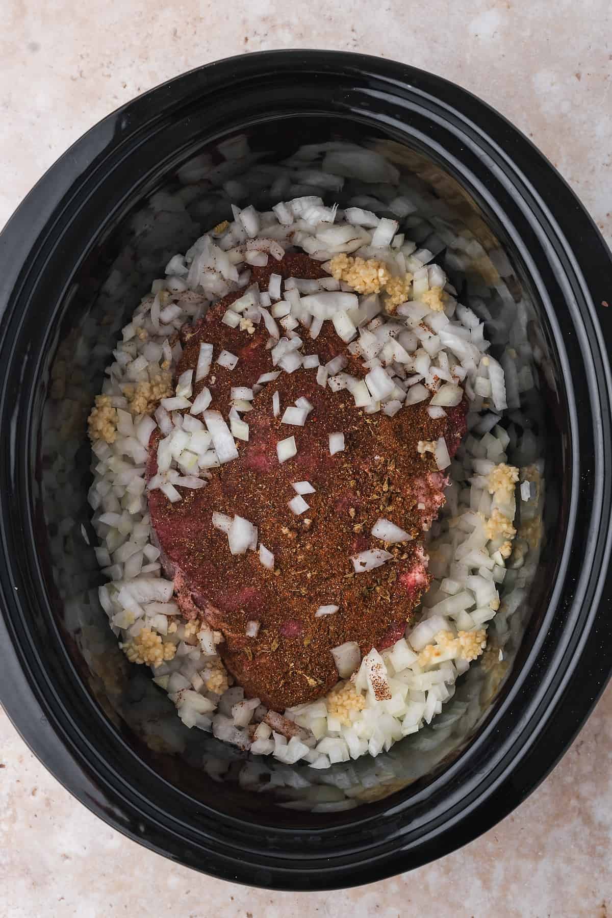 A raw seasoned beef roast placed on top of chopped onions and garlic in a slow cooker, ready for cooking into shredded beef tacos.
