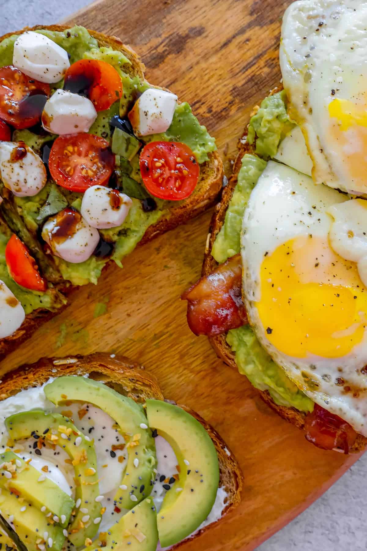Four slices of toast with avocado, bacon, tomatoes and eggs on a wooden cutting board.