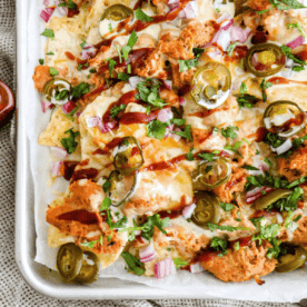 A tray of bbq chicken nachos topped with melted cheese, jalapeños, diced red onions, and fresh cilantro, drizzled with barbecue sauce.