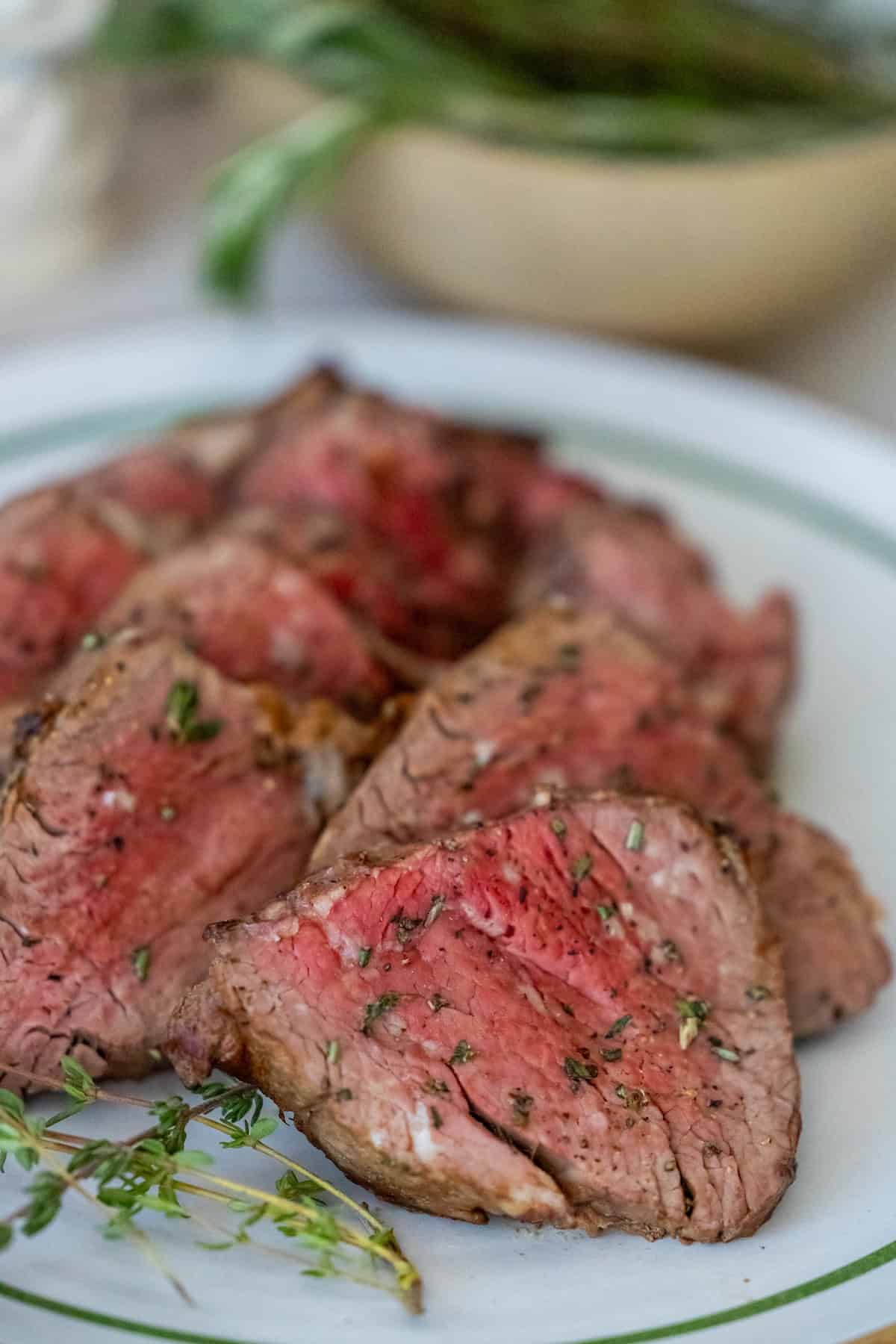 Close-up of sliced, pink-cooked beef tenderloin on a green-rimmed plate, garnished with herbs.