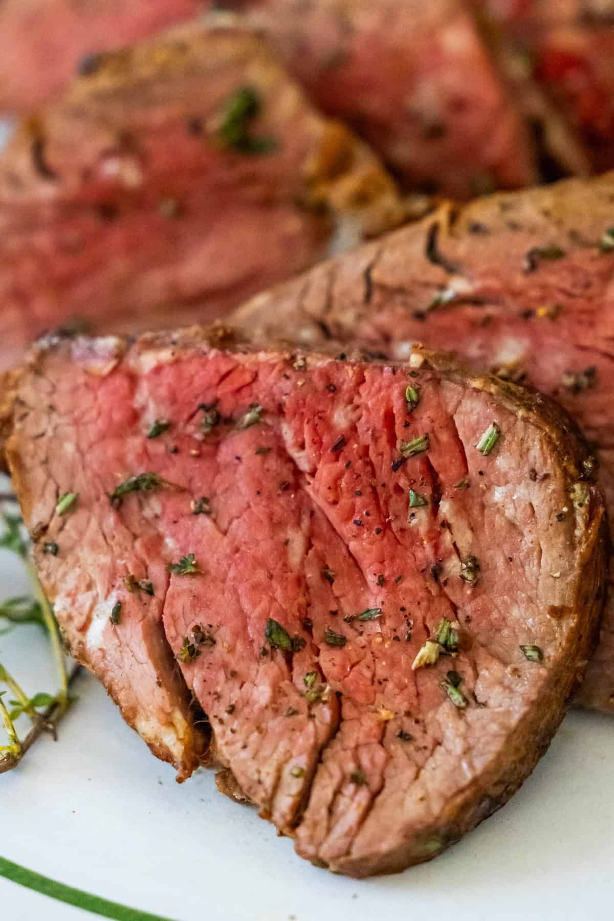 A close-up of sliced medium-rare beef tenderloin, seasoned with herbs, elegantly arranged on a white plate.