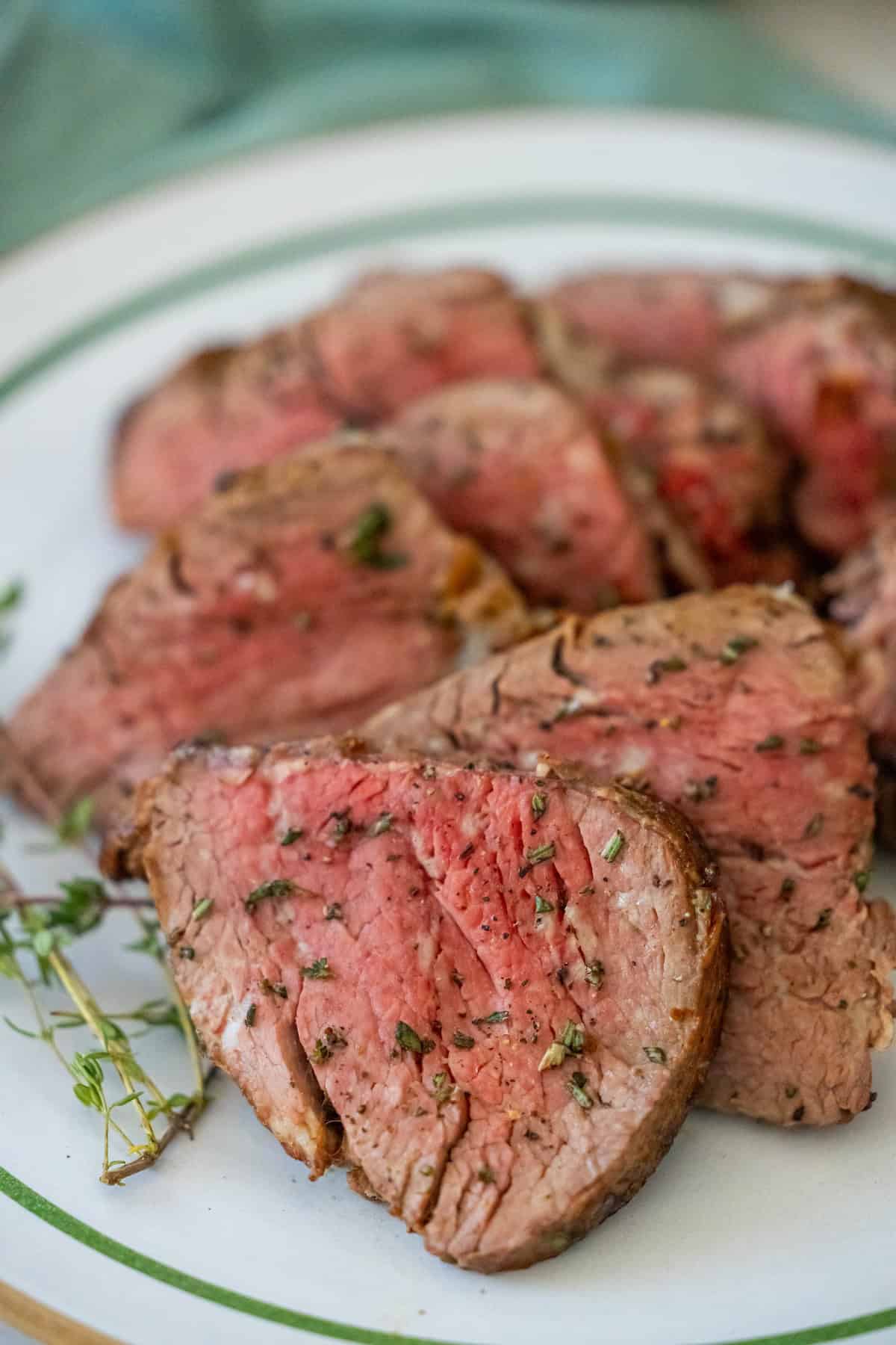 Sliced medium-rare beef tenderloin graces a white plate with green trim, elegantly garnished with fresh herbs.