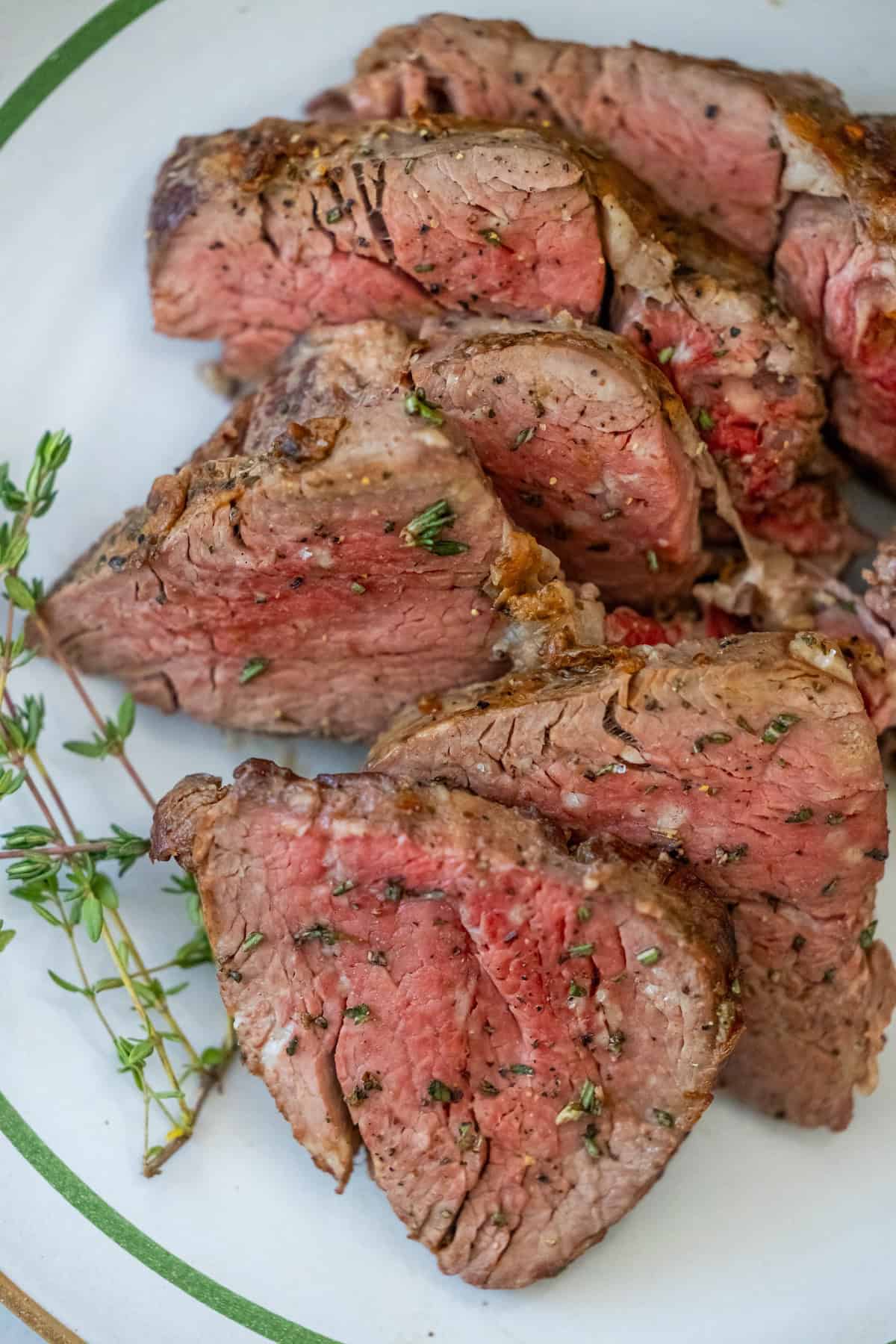 Slices of medium-rare beef tenderloin seasoned with herbs rest on a white plate, elegantly garnished with fresh thyme.