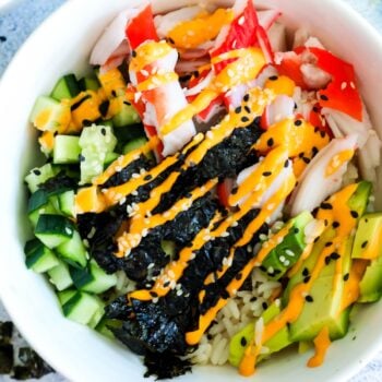 A colorful poke bowl with cucumber, avocado, crab meat, seaweed, and rice, garnished with sesame seeds and spicy mayo.