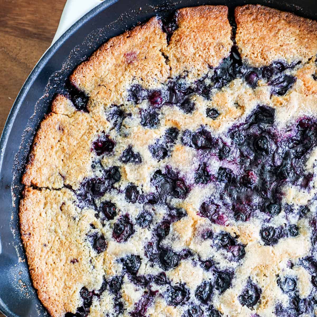 A close-up of a freshly baked blueberry skillet cake, showing a golden-brown crust and juicy blueberries embedded in the surface.