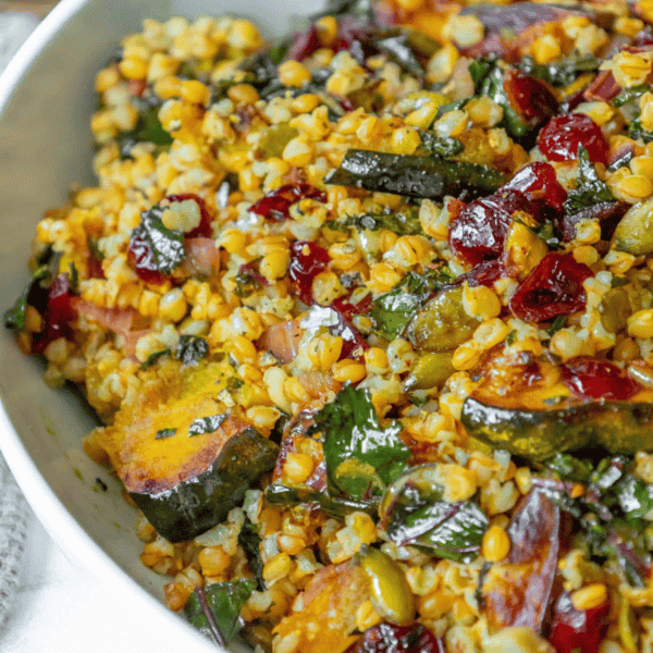 A colorful dish of mixed grains including wheat, with diced vegetables, cranberries, herbs, and acorn squash, served in a white bowl.