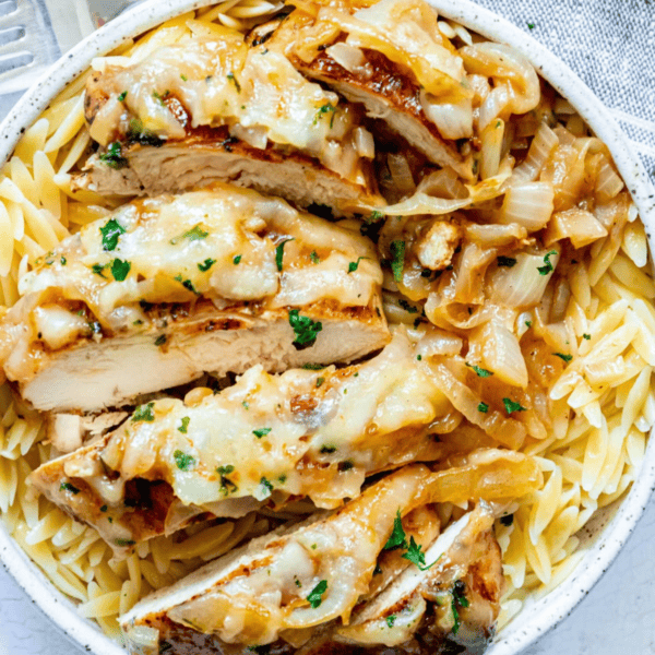 A plate of pasta topped with sliced baked French onion chicken, caramelized onions, and melted cheese, garnished with chopped parsley.