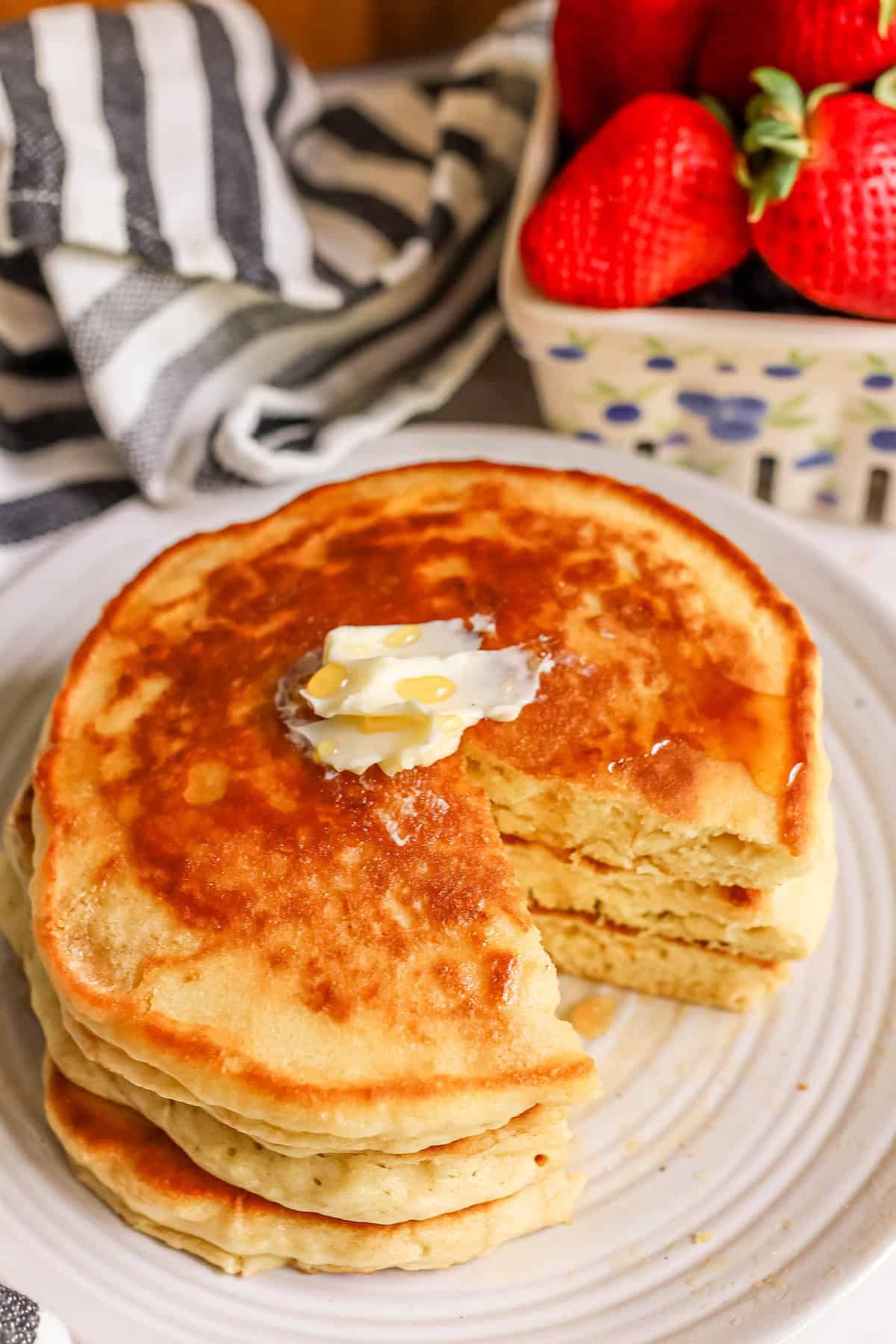 A stack of fluffy pancakes topped with butter and syrup on a white plate, with fresh strawberries in the background.