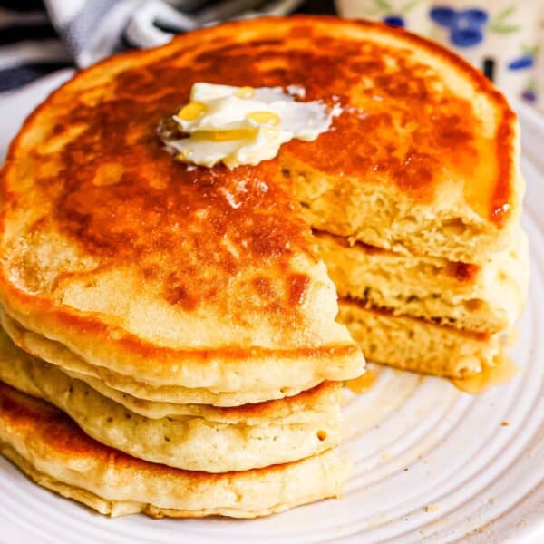 A stack of fluffy pancakes with butter and syrup on a plate, showing a bite taken out of the top pancake.