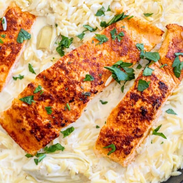 Three pieces of seasoned, grilled salmon are served over a bed of creamy orzo pasta with artichokes, garnished with chopped parsley.
