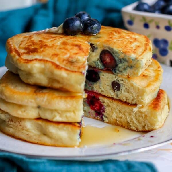 Stack of fluffy pancakes with blueberries, cut open to show berries inside, served on a white plate with a side of extra blueberries.