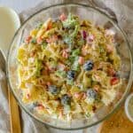 Glass bowl filled with a quick, easy pasta salad, featuring bowtie pasta, black olives, diced red bell peppers, broccoli, and a creamy dressing. The bowl is placed on a beige cloth with a spoon nearby.