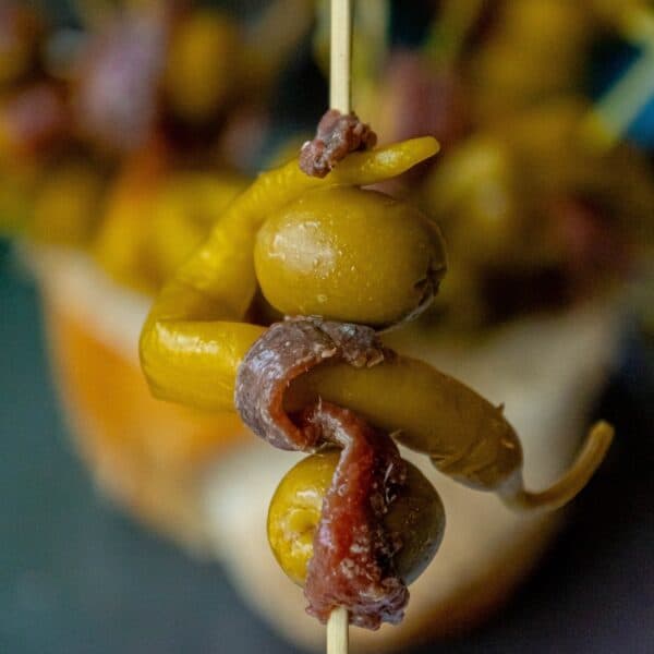 Close-up of a Gilda skewer with green olives, a pickled pepper, and an anchovy slice wrapped around the ingredients. The background shows a blurred view of similar pintxos.