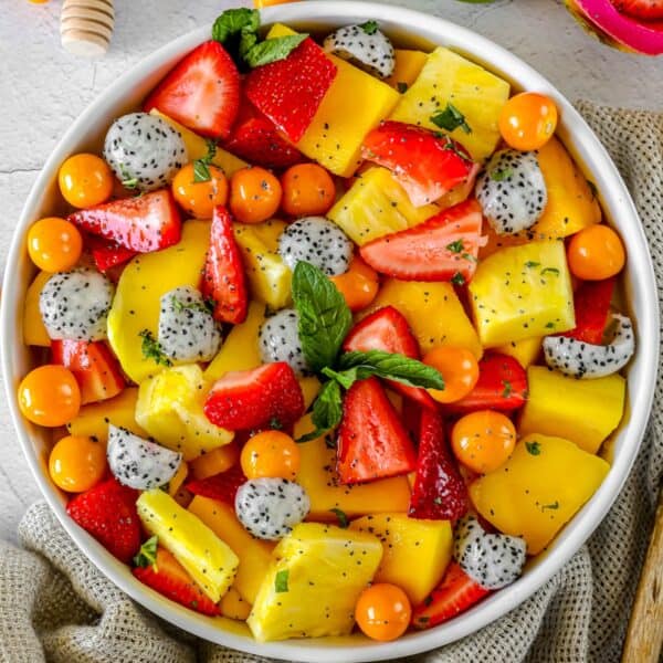 A bowl filled with a colorful tropical fruit salad consisting of sliced strawberries, pineapple, mango, dragon fruit, and golden berries, garnished with fresh mint leaves.