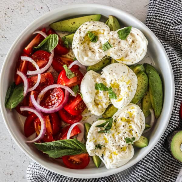 A bowl of burrata tomato and avocado salad with sliced red onions, topped with basil and olive oil.