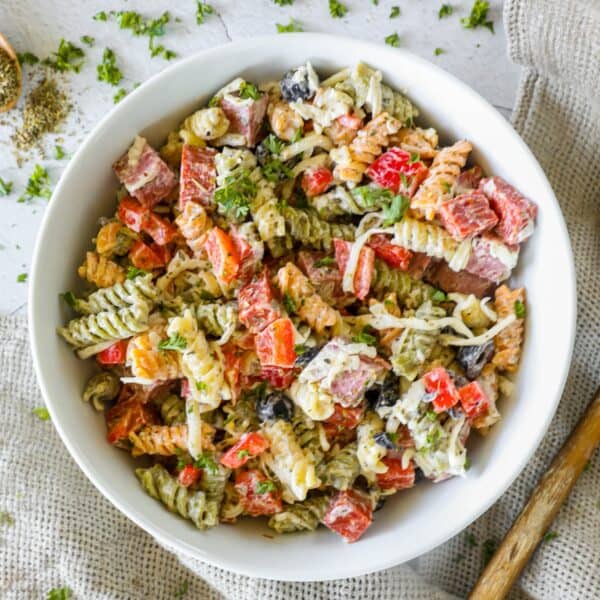 A bowl of the best Italian pasta salad featuring tricolor rotini mixed with diced red bell peppers, black olives, chopped parsley, and shredded cheese, set on a textured cloth background.