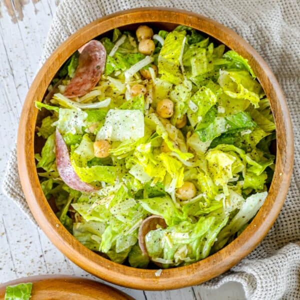 A wooden bowl filled with a green salad, reminiscent of a La Scala Chopped Salad, including lettuce, chickpeas, shredded cheese, and slices of cured meat, placed on a textured napkin.