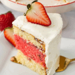 Slice of layered strawberries and cream cake with white frosting, garnished with a halved strawberry on top, positioned on a white plate with a gold fork beside it.