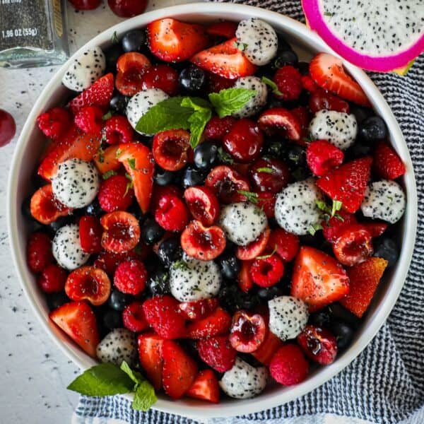 A white bowl filled with a colorful mix of strawberries, blueberries, raspberries, cherries, and sliced dragon fruit, garnished with fresh mint leaves, placed on a blue and white cloth.