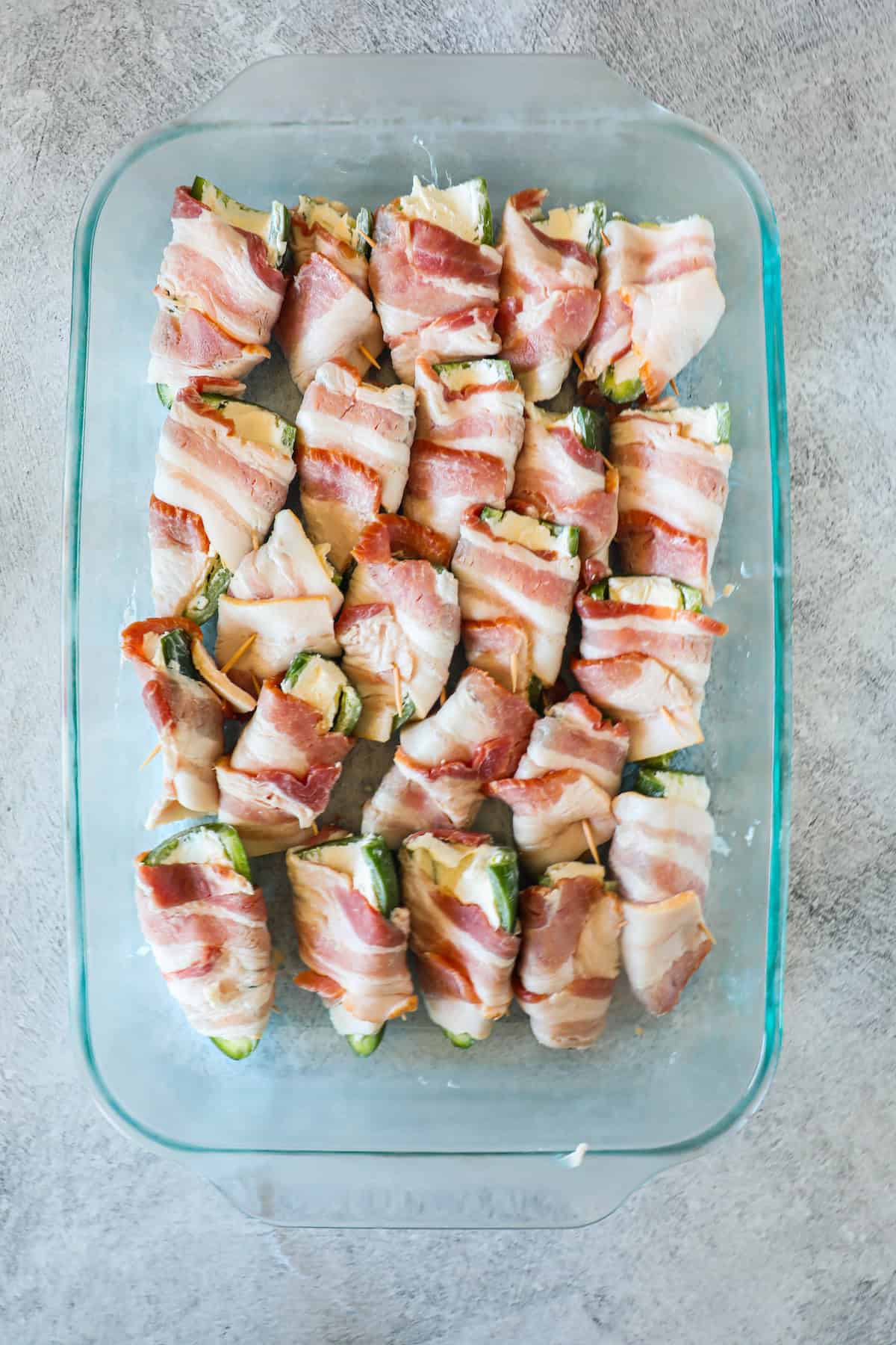 A glass baking dish filled with bacon-wrapped jalapeรฑo poppers, arranged in rows on a gray surface.