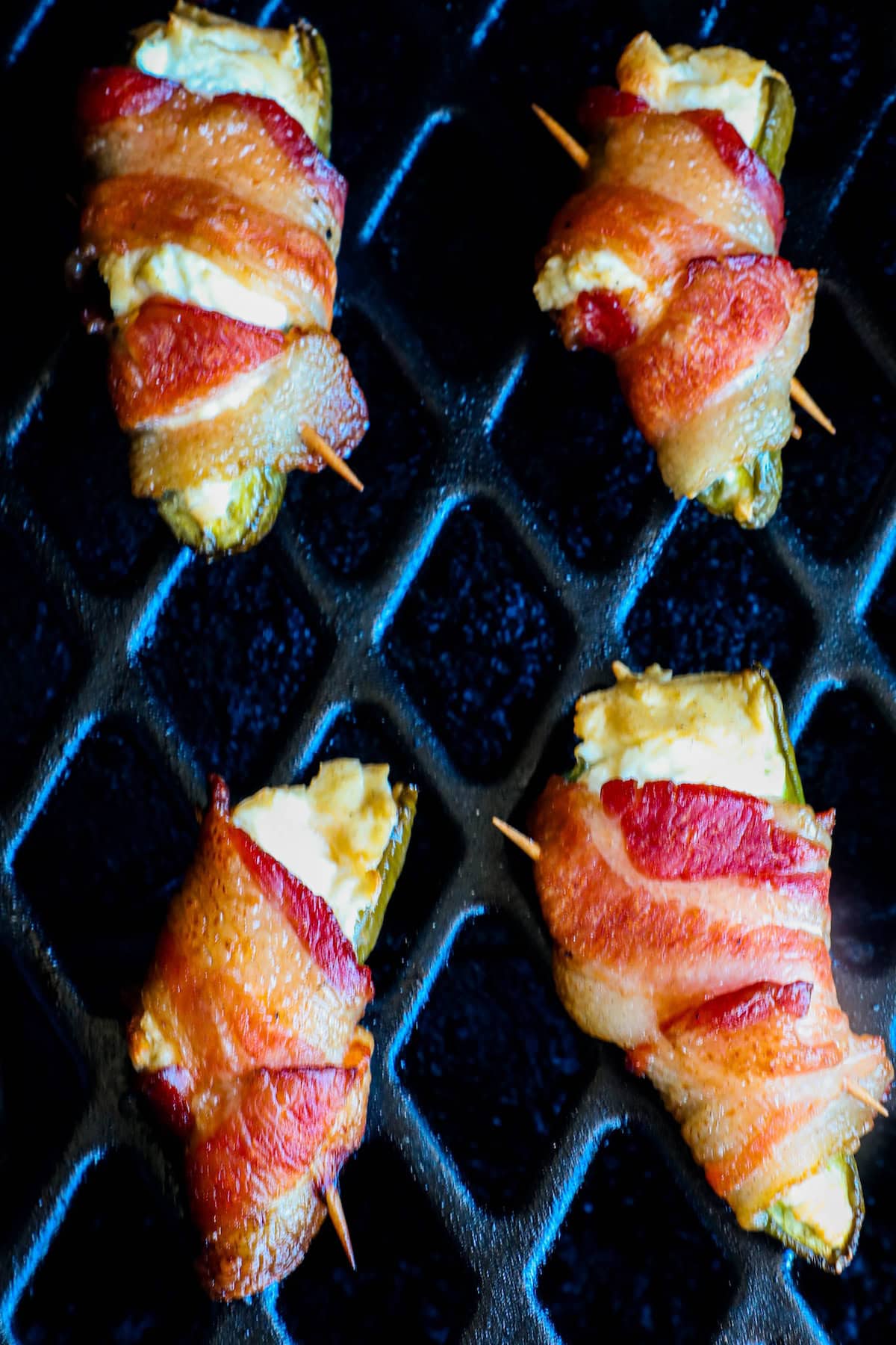 Four bacon-wrapped jalapeรฑo poppers on a black grill.