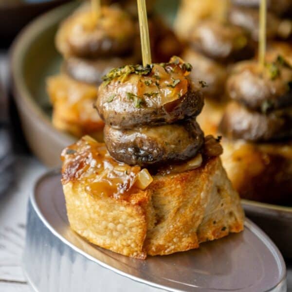 Close-up of a skewer with two mushrooms on a piece of bread, topped with cooked onions and herbs, placed on a metal tin. A perfect example of Pintxos de Txampis. More skewers are visible blurred in the background.