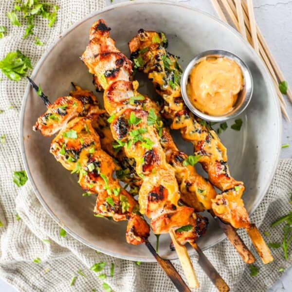 A bowl of grilled chicken skewers garnished with herbs, served with a bowl of dipping sauce on a light-colored napkin.