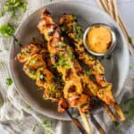 A plate with grilled chicken skewers garnished with herbs, accompanied by a small dish of dipping sauce.