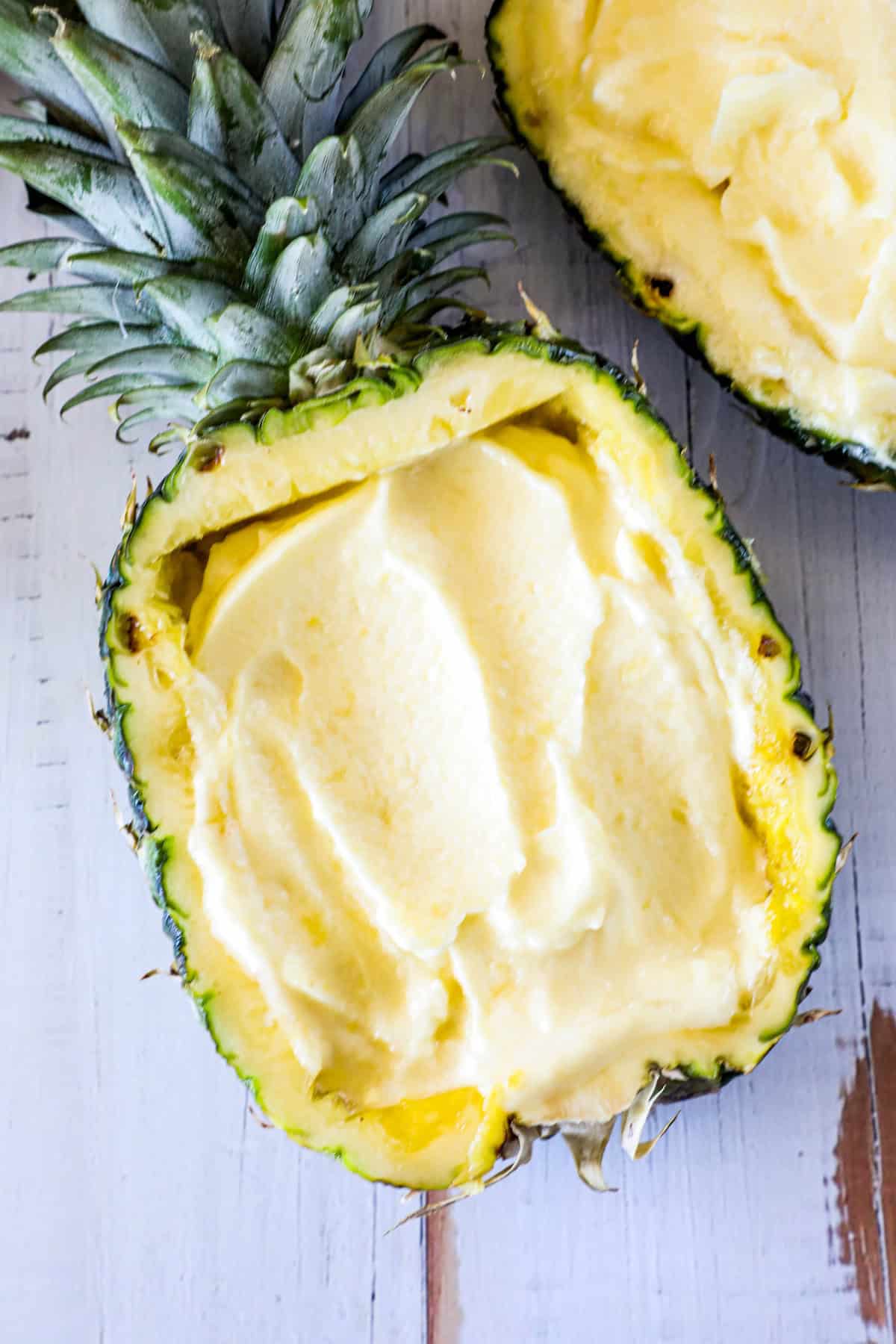 A hollowed-out pineapple half filled with a creamy yellow dessert, placed on a light-colored wooden surface.