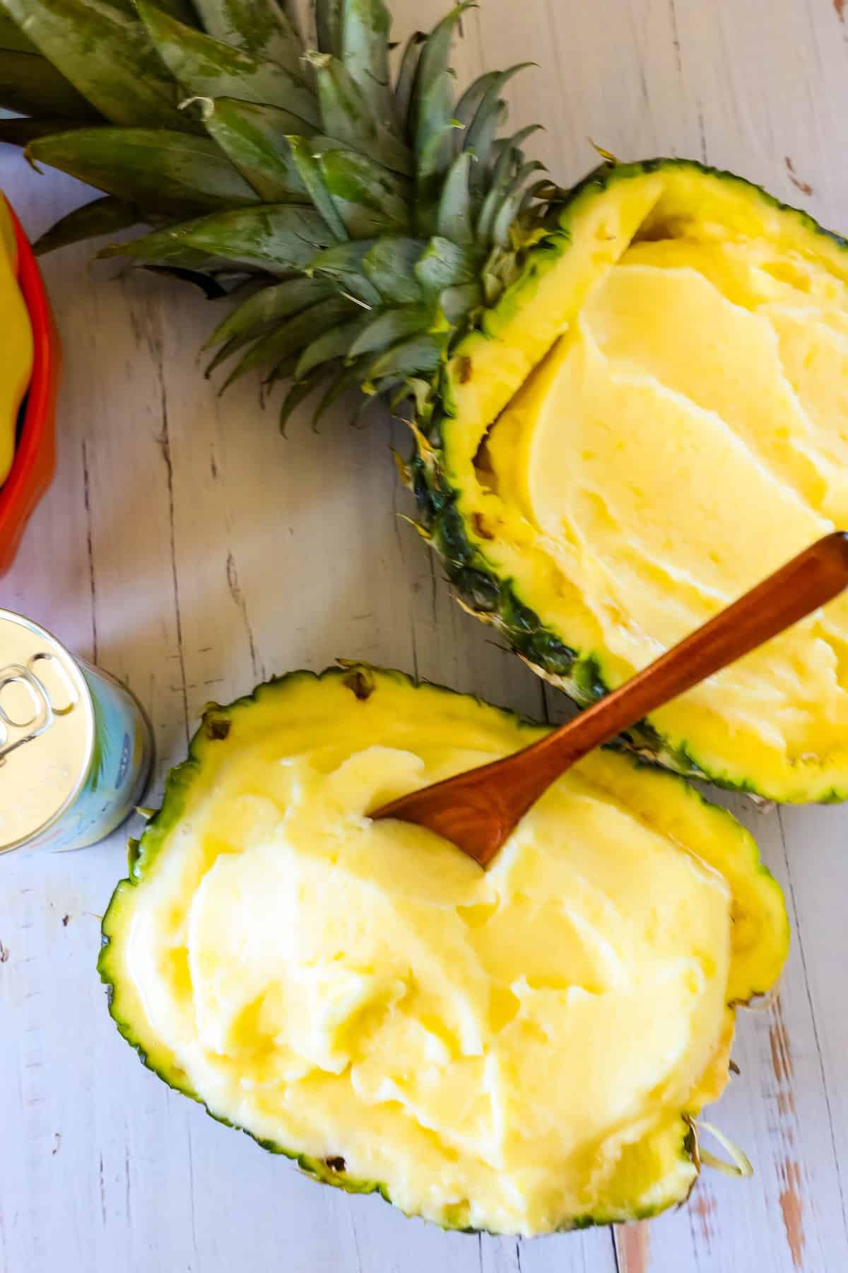 A halved pineapple filled with pineapple soft serve, with a wooden spoon placed in one half, and a can nearby on a wooden surface.