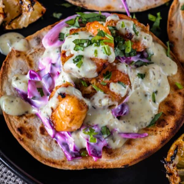 A grilled taco topped with shrimp, purple cabbage, creamy sauce, and chopped green herbs, served on a black plate.