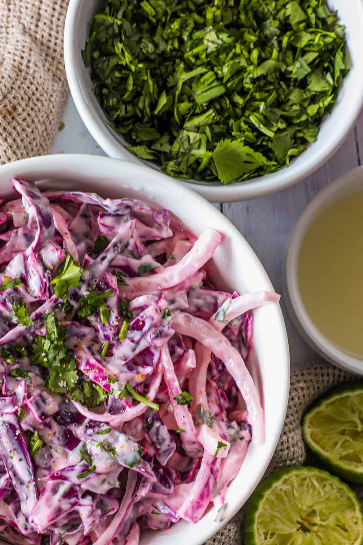 A bowl of creamy coleslaw made with purple cabbage and garnished with herbs, alongside a bowl of chopped herbs, lime halves, and a small bowl of dressing.