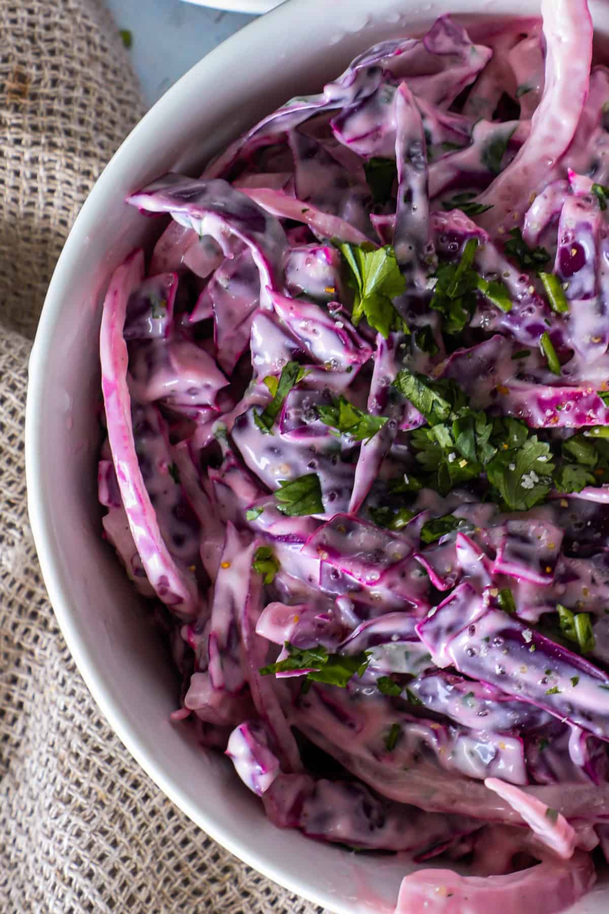 Close-up of a bowl of creamy coleslaw with shredded purple cabbage mixed with a white dressing and garnished with chopped green herbs. The bowl is placed on a rustic fabric surface.