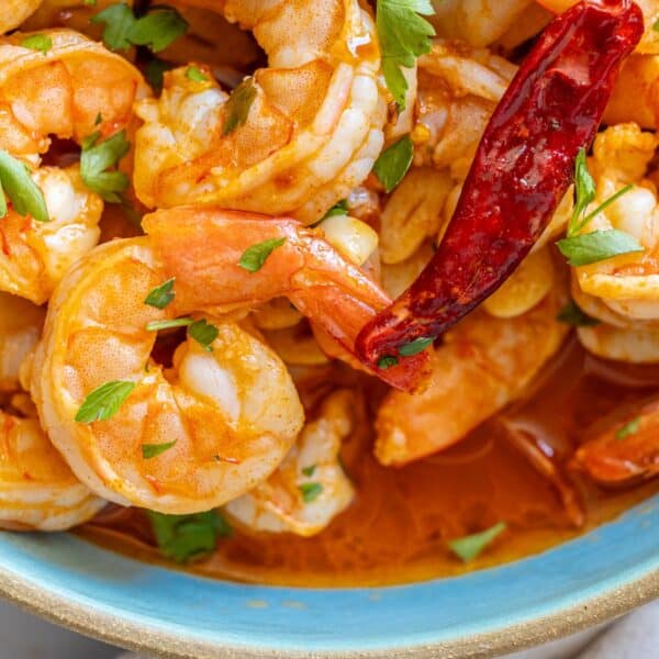 Close-up of a bowl of shrimp in a spicy red sauce, bursting with garlic and garnished with a whole dried chili pepper and fresh parsley.