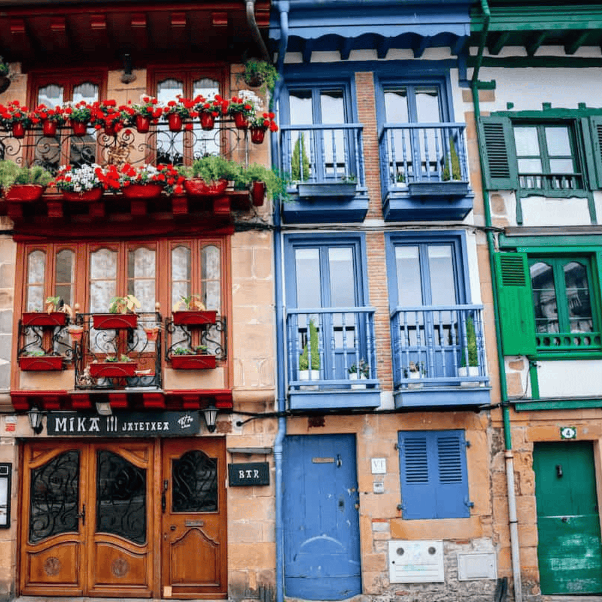 Colorful row of houses with red, blue, and green facades; features balconies and flowers. The ground floor includes a restaurant with the sign "MIKA Jatetxea." Perfect for any travel guide to Hondarribia.