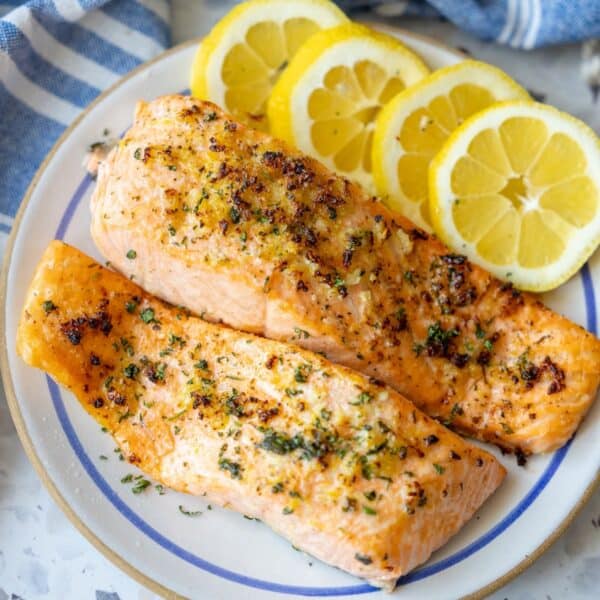 Two grilled salmon fillets garnished with herbs are served on a plate with three slices of fresh lemon, offering a delicious twist inspired by the Easy Air Fryer Lemon Garlic Salmon Recipe.